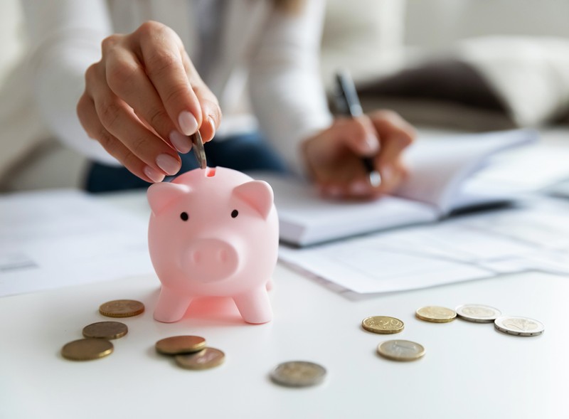 Focused on her hands, a woman, holds a pen and writes in a notebook while placing a coin in a piggybank. woman puts coins in piggybank
