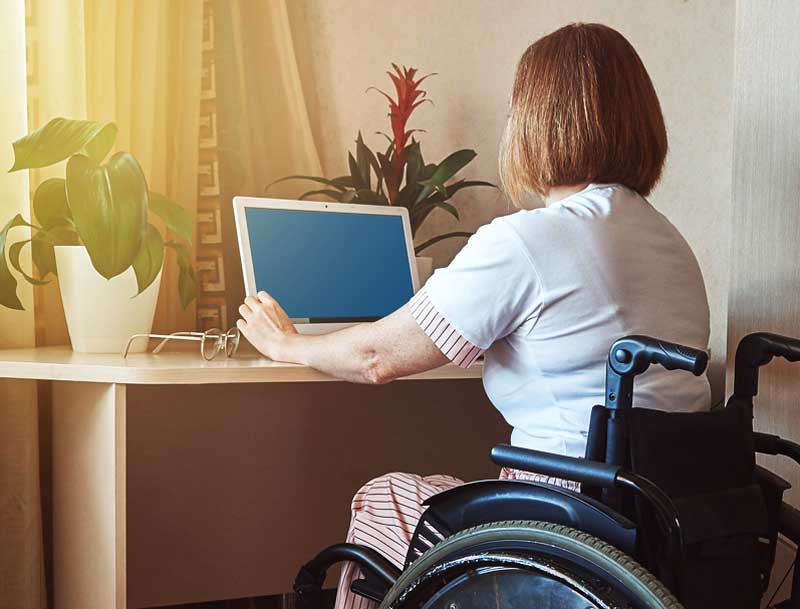 A disabled Veteran sits in a wheelchair at her desk, starting up a tablet. There are spectacles and plants on her desk. a disabled veteran in wheelchair turns on a tablet at her desk