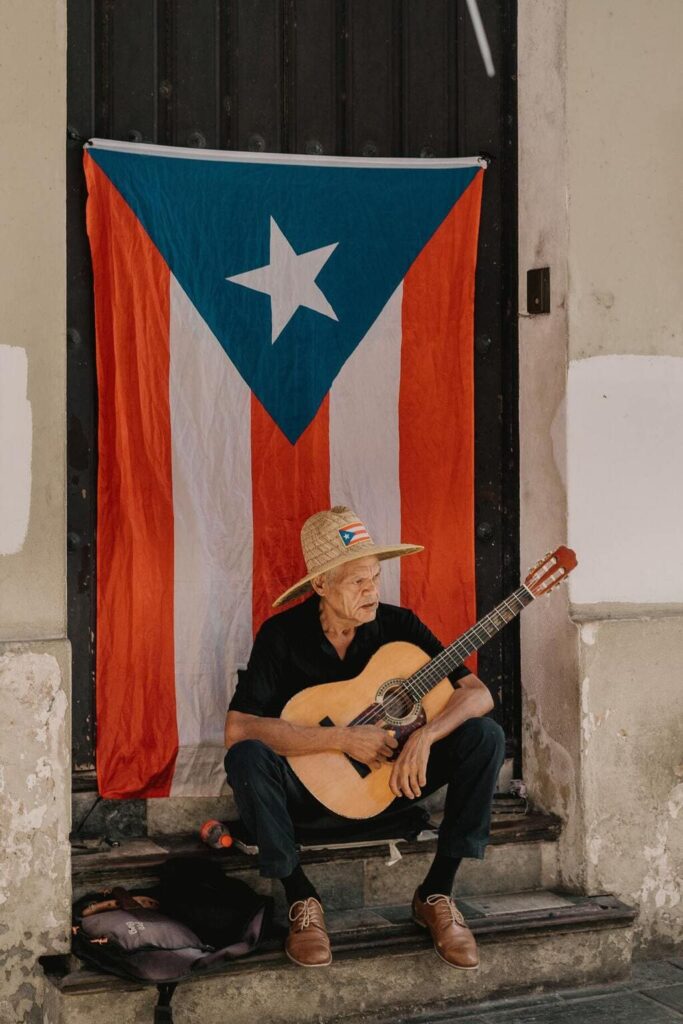 Older man in straw hat holding a guitar in front of a Puerto Rico flag on the streets of Old San Juan.